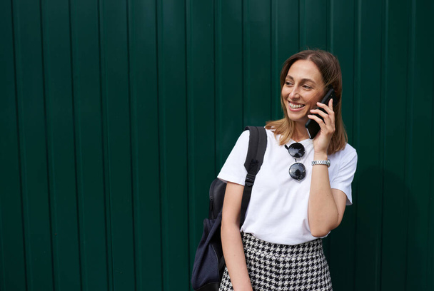 Outdoor image of happy young woman in white t-shirt with black sunglasses and backpack talking on mobile phone, standing against green wall. Female student waiting for a colleague to going to college - Foto, afbeelding