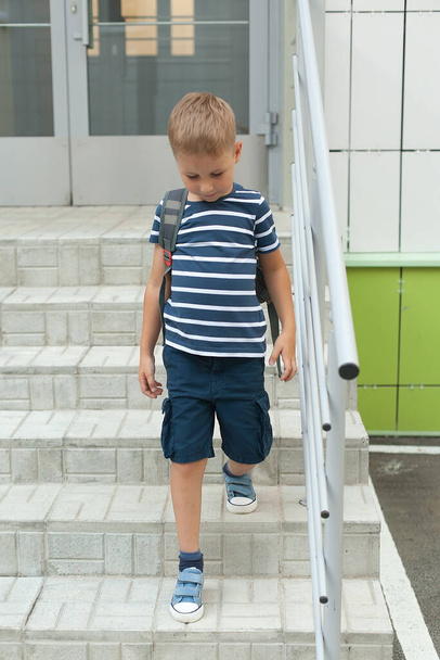 Ένα μικρό αγόρι περπατάει μόνο του, φεύγει από το κτίριο και κατεβαίνει τα σκαλιά. Νηπιαγωγείο. - Φωτογραφία, εικόνα