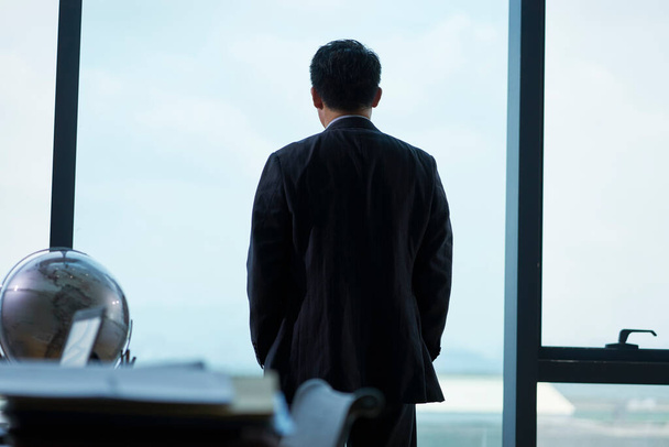 πίσω όψη ενός ασιατικού εταιρικού στελέχους στέκεται από το παράθυρο στο γραφείο κοιτάζοντας έξω - Φωτογραφία, εικόνα