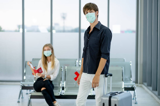 Портрет красивого белого мужчины, стоящего перед женщиной, сидит на стуле в аэропорту во время снятия изоляции от пандемии Ковид-19 по всему миру. - Фото, изображение