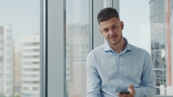 Портрет бізнесмена, який посміхається і читає повідомлення по телефону, в офісі з панорамними вікнами
. - Кадри, відео