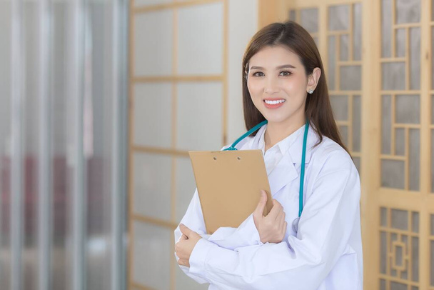 Молодая красивая азиатка-врач, стоящая со скрещенными руками счастливая и улыбающаяся в больнице. В белом халате и стетоскопе. - Фото, изображение