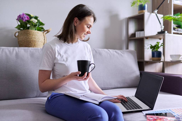 Young woman with laptop computer, cup of coffee sitting on couch, home - Foto, Imagen