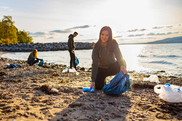 Χαμογελαστή νεαρή γυναίκα καθαρισμού παραλία με εθελοντές κατά τη διάρκεια του ηλιοβασιλέματος - Φωτογραφία, εικόνα