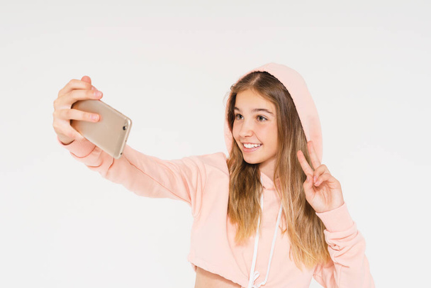 Cute smiling teen girl in pink hoodie taking selfie on smartphone isolated on white background - Photo, Image