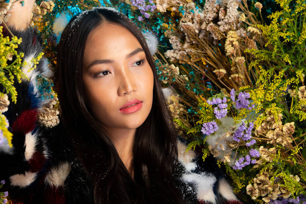 Beautiful romantic young Asian woman in Black Fur cloth in bush variety of flowers posing on background fresh and dried flora. Inspiration of autumn winter snow Perfume, cosmetics concept. - Foto, afbeelding