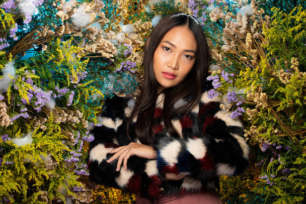 Beautiful romantic young Asian woman in Black Fur cloth in bush variety of flowers posing on background fresh and dried flora. Inspiration of autumn winter snow Perfume, cosmetics concept. - 写真・画像