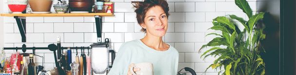 Портрет горячие волосы красивая женщина в повседневной одежде в черной кухне. Женщина держит чашку горячего чая кофе Фото расслабляющий уютный интерьер Длинный баннер - Фото, изображение