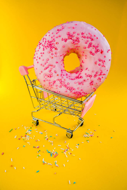 黄色の背景のショッピングカートで甘いピンクのドーナツ. - 写真・画像