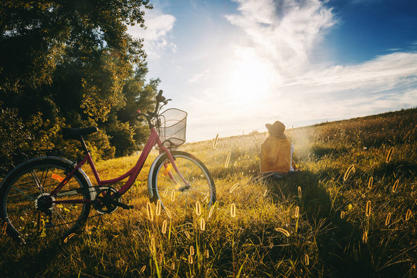 Woman in a hat sits on the grass looking at the forest at sunset. The bike stands in golden grass with spikelets. Beautiful landscape - Photo, Image