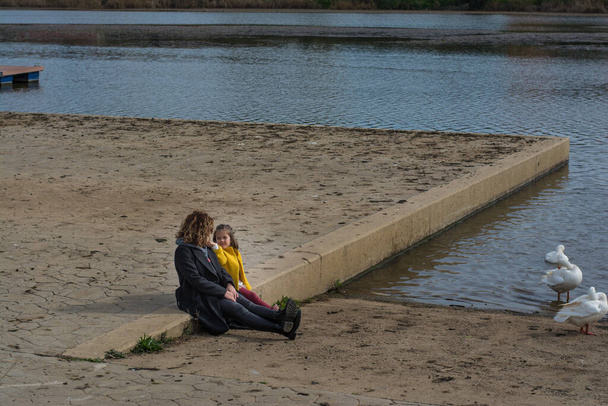 Nainen tyttärensä kanssa laiturilla katsellessaan jokea. Nainen ja tyttö istumassa laiturilla katsomassa ankkoja joessa. Perhekäsite. Onnellinen äiti ja hänen tyttärensä pitää hauskaa.  - Valokuva, kuva