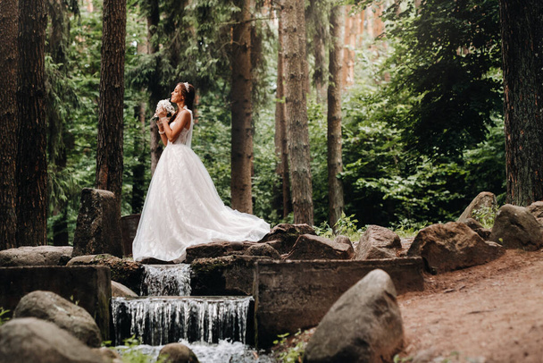 An elegant bride in a white dress and gloves holding a bouquet stands by a stream in the forest, enjoying nature.A model in a wedding dress and gloves in a nature Park.Belarus. - Fotoğraf, Görsel
