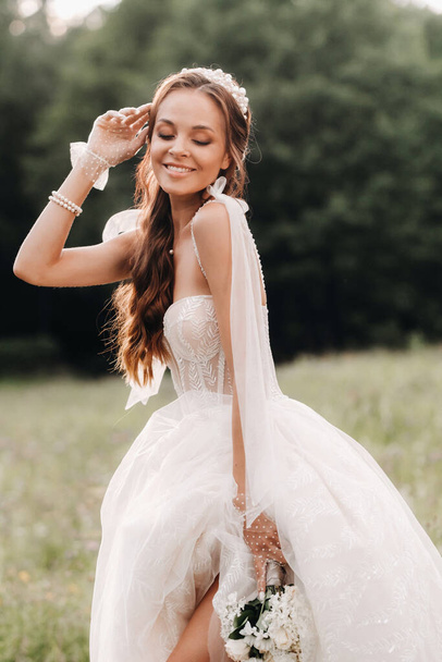 On the wedding day, an Elegant bride in a white long dress and gloves with a bouquet in her hands stands in a clearing enjoying nature. Belarus - Foto, imagen