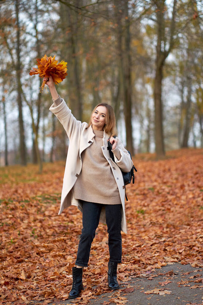 Moderne jeugdcollectie. Jong blond vrolijk meisje met trui, zwarte jeans, trendy herfstjas en leren zwarte laarzen met boeket herfstbladeren. Herfst park op achtergrond.                               - Foto, afbeelding