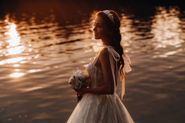An elegant bride in a white dress and gloves stands by the river in the Park with a bouquet, enjoying nature at sunset.A model in a wedding dress and gloves in a nature Park.Belarus. - Photo, image