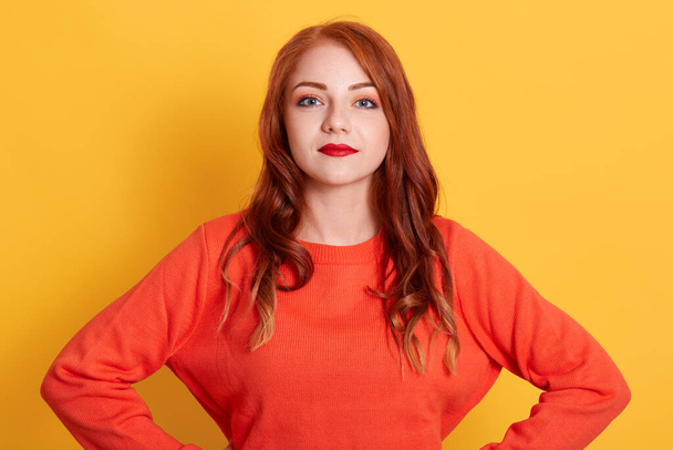 Gorgeous red haired young European lady in orange casual sweater looking at camera with calm facial expression, posing with red lips isolated over yellow wall. - 写真・画像