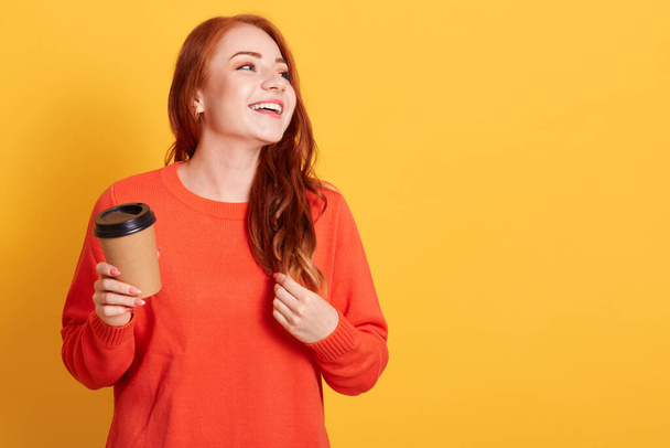 Young Caucasian woman over yellow background holding coffee to take away, laughing and looking aside, copy space for advertisement or promotional text. - Photo, image