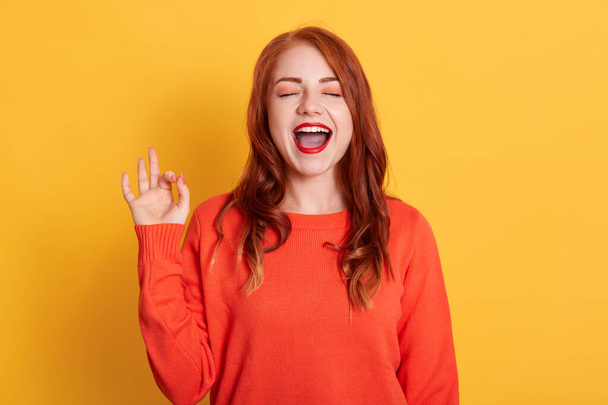 Young red haired beautiful female with closed eyes, showing ok sign and screaming, posing isolated over yellow background, lady wearing orange casual sweater. - Фото, зображення
