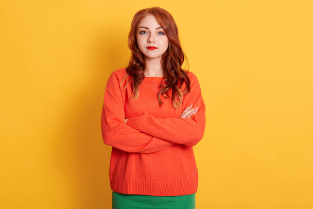 Portrait of young tender red haired european woman with serious look, wearing orange sweater, looking at camera with calm or sad expression, caucasian female model posing with folded hands. - Zdjęcie, obraz