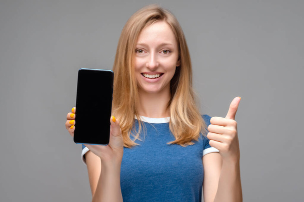 jong mooi blond vrouw tonen scherm van smartphone over geïsoleerde achtergrond blij met grote glimlach doen OK teken, duim omhoog met vingers, uitstekend teken - Foto, afbeelding