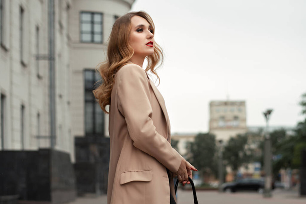 Jeune belle femme avec de longs cheveux blonds en trench-coat beige et sac posant sur la rue de la ville d'automne. Concept de style de vie - Photo, image