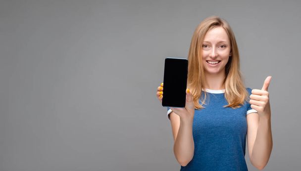 Young beautiful blonde woman showing screen of smartphone over isolated background happy with big smile doing OK sign, thumb up with fingers, excellent sign. Copy space for your text - Valokuva, kuva