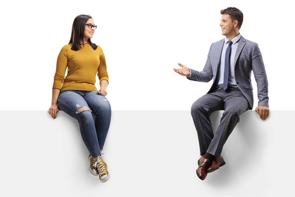 Young woman with glasses sitting on a blank panel and talking to a man in a suit isolated on white background - Φωτογραφία, εικόνα
