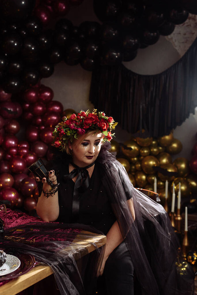 Gyönyörű nő elegáns fekete ruhában egy pohár borral ünnepli születésnapját, halloween. Egy felnőtt boszorkány egy Halloween-re berendezett szobában.. - Fotó, kép