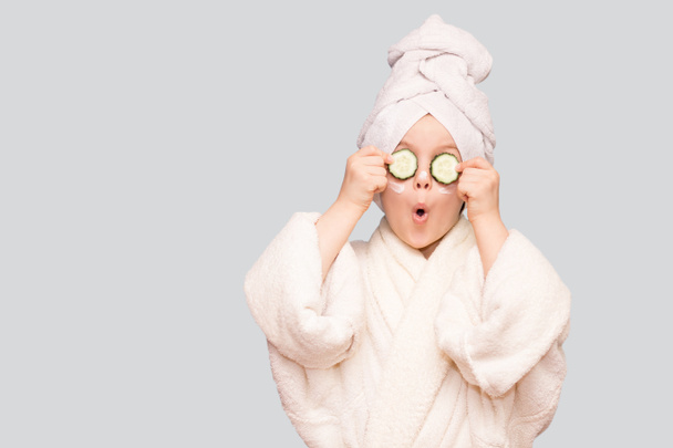 Funny little girl in white coat and towel with mask for skin face and cucumbers on eyes isolated on white background. Spa skin care treatments - Photo, Image