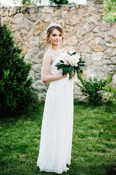Stylish happy smile bride with bouquet of peonies with crown, stand, poses on grass on background in courtyard. Close up. Nature. Against the background of the stone wall. Looking forward. - Foto, afbeelding