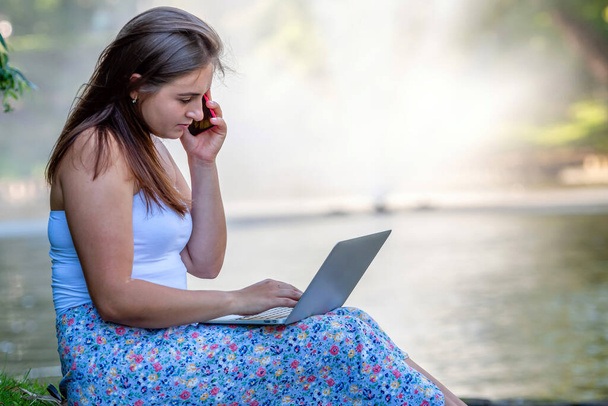 a young woman sits on city park near the canal, talking on the phone and working on a laptop, remote training and work concept - Photo, Image