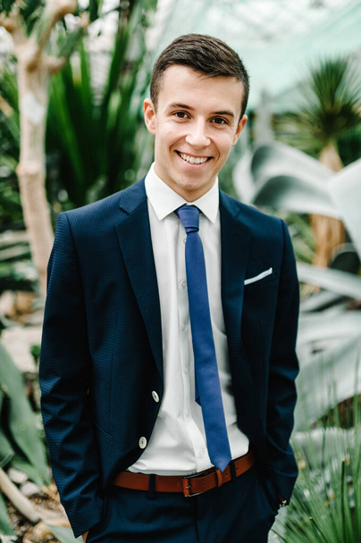Happy portrait handsome groom in a wedding suit and tie is standing on the background of greenery. Man stands in the Botanical garden full of greenery. - Foto, imagen