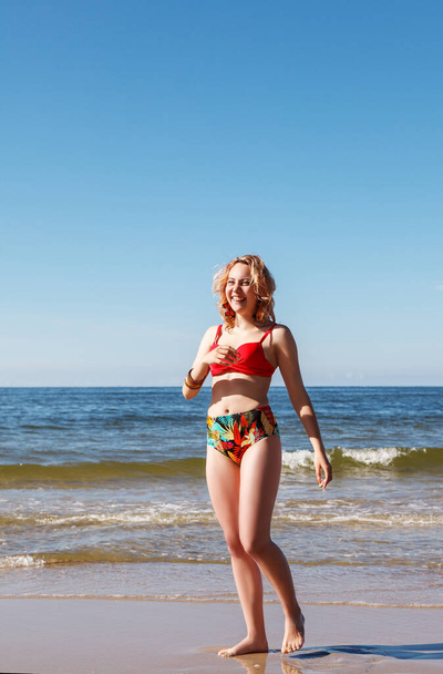 young blonde smiling girl in a red swimsuit posing standing on the seashore on sunny summer day - Фото, изображение