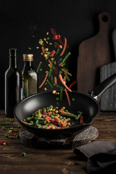 Frozen vegetables: a mixture of vegetables, string beans and cauliflower is poured into a black pan on a dark background with bottles of oil and cutting boards. Flying food close-up with a copyspace. Vertical orientation. - Foto, afbeelding