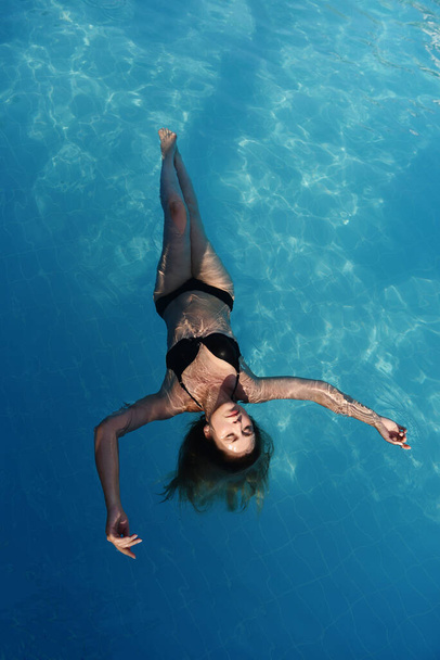 Высокоугольный вид привлекательной женщины, отдыхающей в воде с закрытыми глазами. Портрет счастливой женщины в бикини, плавающей в воде. - Фото, изображение