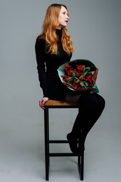 Portrait of red-haired young girl florist sitting on chair with bouquet of flowers in emerald package on light background. Rose, ilex, spruce. - Фото, изображение