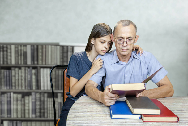 Granddaughter and grandfather read a book. They sit at the table, in the room. Next to the bookcase. Books on the table. Grandfather opened the book, granddaughter looks at the book. - Photo, Image