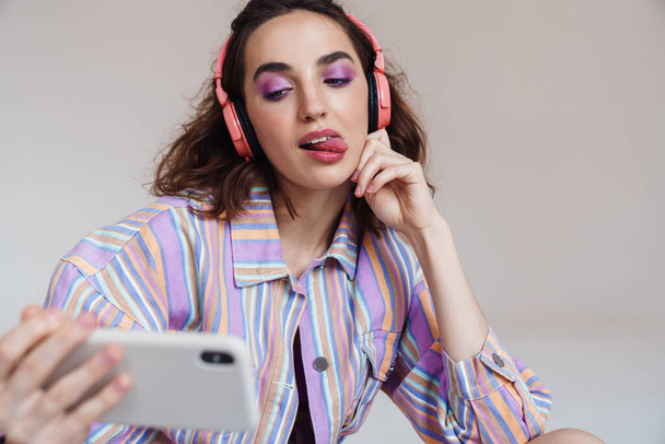 Image of amusing woman showing her tongue while using cellphone and headphones isolated over white wall - Фото, изображение