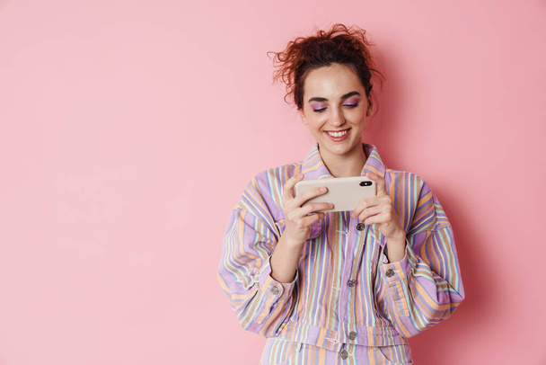Image of ginger smiling woman playing video game on cellphone isolated over pink background - Photo, Image