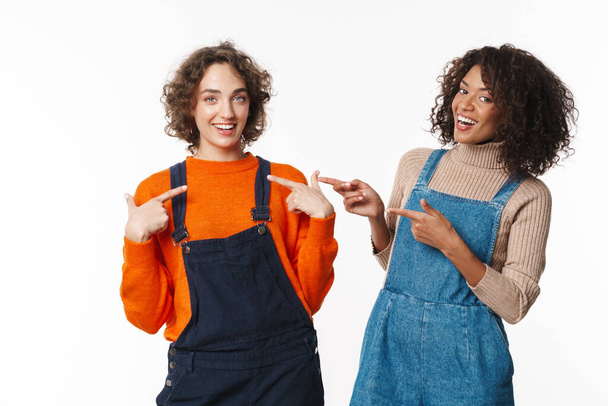Portrait of two joyful women in overalls smiling and pointing fingers isolated over white background - Photo, Image