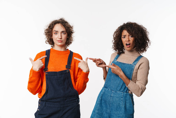 Portrait of two confused women in overalls looking at camera and pointing fingers isolated over white background - Foto, Imagem