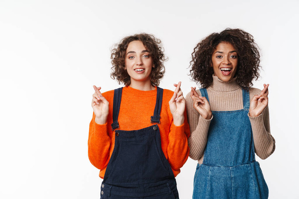 Portrait of happy multinational women in overalls looking at camera with fingers crossed isolated over white background - Фото, зображення