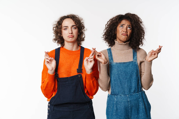 Portrait of unhappy multinational women in overalls looking at camera with fingers crossed isolated over white background - Φωτογραφία, εικόνα