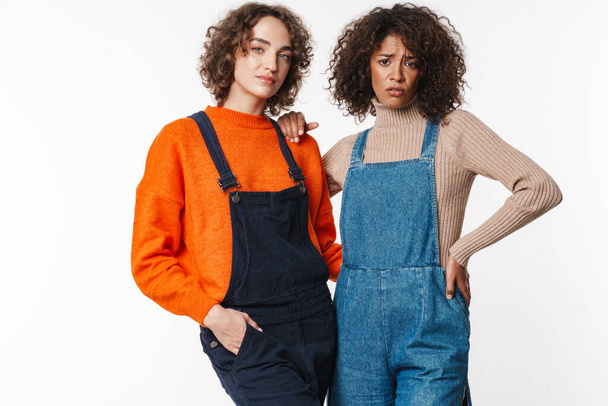Portrait of confused multinational women in overalls looking at camera and hugging isolated over white background - Foto, immagini