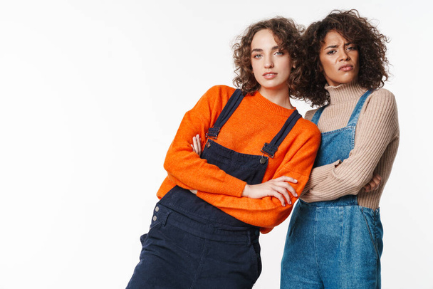 Portrait of confused multinational women in overalls looking at camera with arms crossed isolated over white background - Zdjęcie, obraz