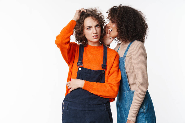 Portrait of african american woman in overall whispering to confused girl isolated over white background - Foto, Imagen