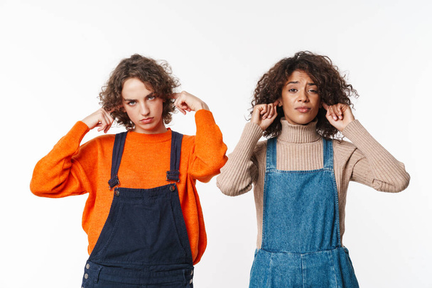 Portrait of serious multinational women in overalls plugging their ears and looking at camera isolated over white background - Zdjęcie, obraz