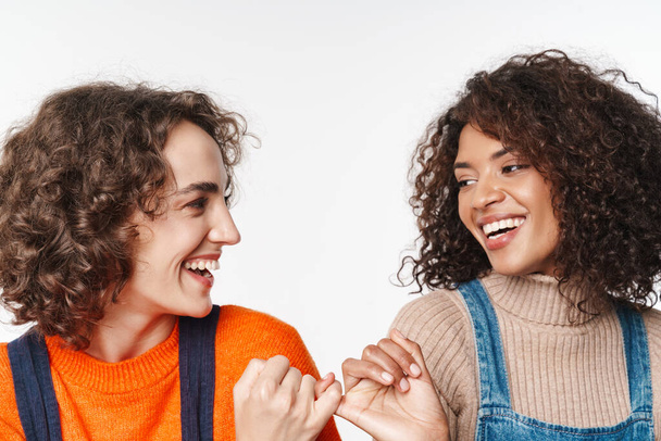 Image of a beautiful optimistic two multiracial girls friends in denim jumpsuits isolated over white wall background holding little fingers of each other. - Photo, Image