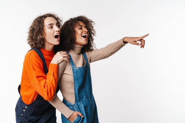 Portrait of surprised multinational women in overalls pointing fingers aside isolated over white background - Zdjęcie, obraz