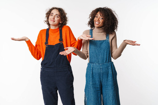 Portrait of cute multinational women in overalls looking at camera and throwing up hands isolated over white background - Foto, Bild